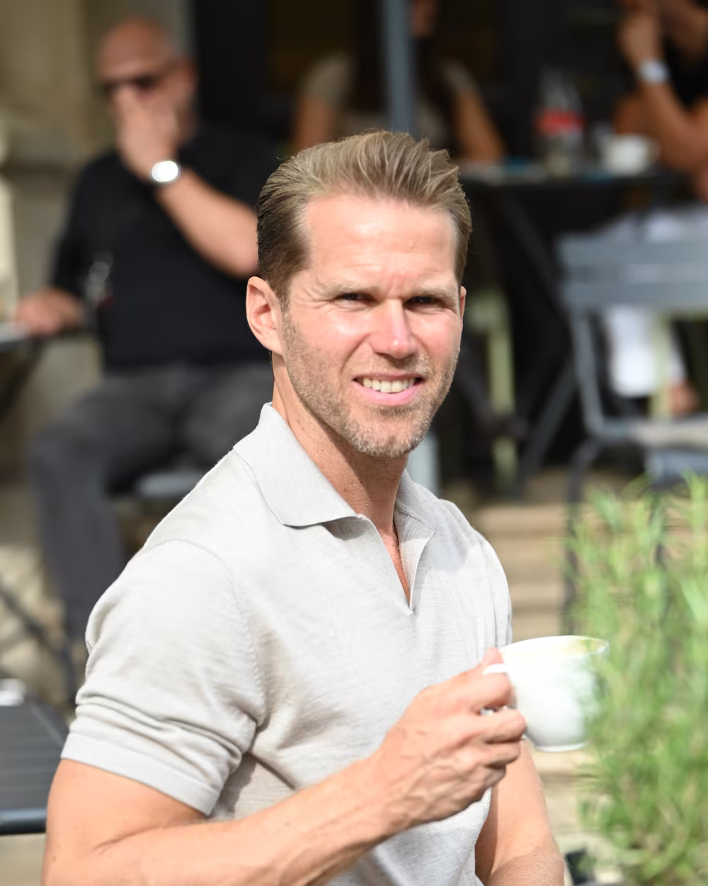 Norman Bartsch im hellen Polo mit einer Tasse Kaffee, lächelnd.