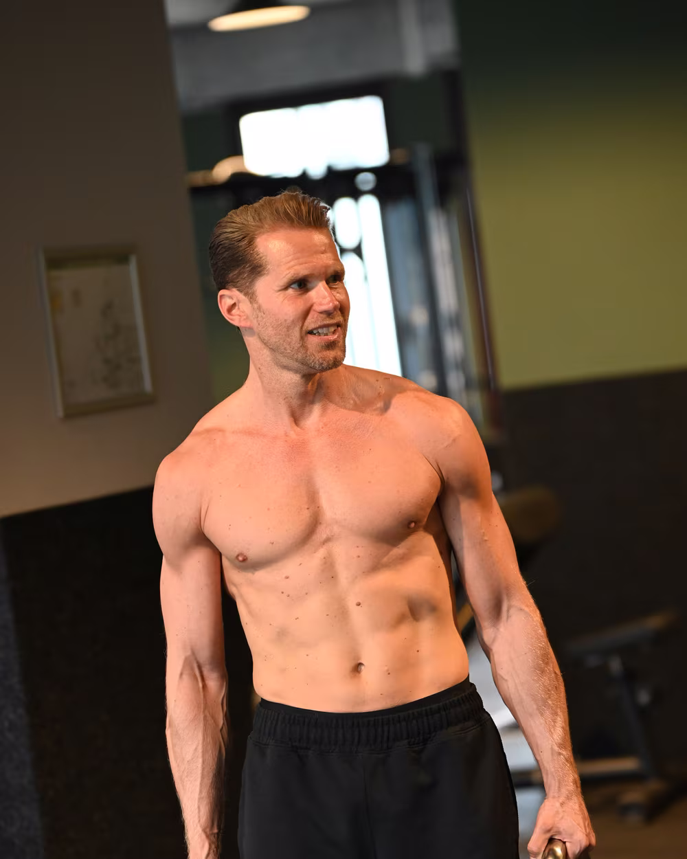 Norman Bartsch mit freiem Oberkörper im Fitnessstudio, die Kettlebells in den Händen.