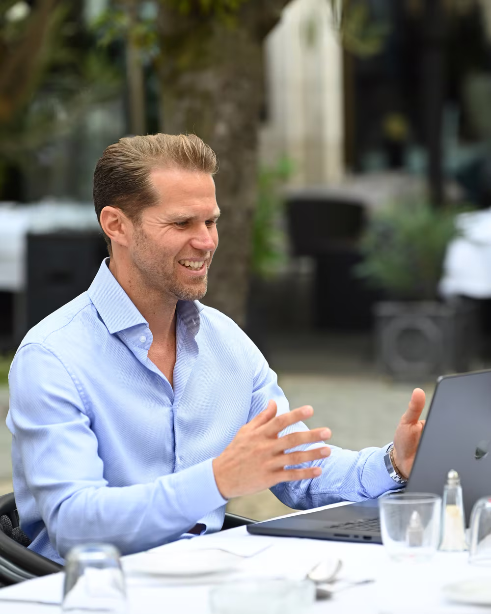 Norman Bartsch am Laptop in einem Straßencafé, im Gespräch mit beiden Händen.